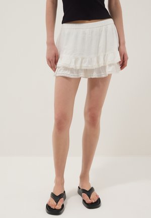 Mujer con falda mini blanca de volantes con ribete de encaje, top negro sin mangas y sandalias negras de plataforma con tira entre los dedos, de pie sobre un suelo blanco.