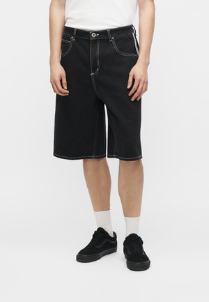 Άτομο που φοράει μαύρα τζιν σορτς μέχρι το γόνατο με λευκή αντίθετη ραφή, λευκές κάλτσες, μαύρα αθλητικά παπούτσια και λευκό μπλουζάκι.