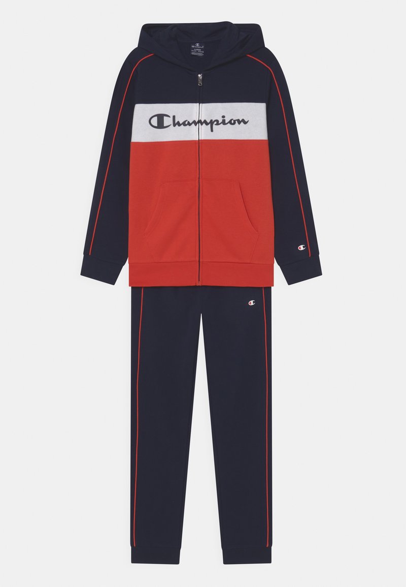 Tuta rossa e blu con giacca a zip con cappuccio caratterizzata da una striscia grigia e logo "Champion", abbinata a pantaloni coordinati e dettagli accentuati.