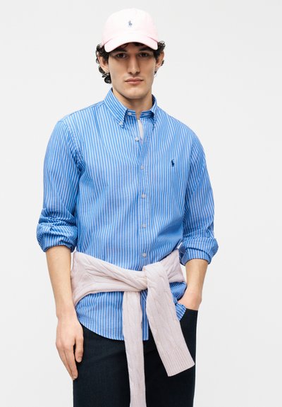 Νεαρός άνδρας που φοράει μπλε και λευκό ριγέ πουκάμισο με κουμπιά, ροζ καπέλο μπέιζμπολ και ανοιχτό ροζ πουλόβερ δεμένο γύρω από τη μέση του.