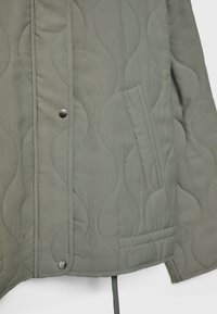 Gros plan sur le devant d’une veste matelassée verte montrant des boutons-pression, une poche latérale diagonale et un motif de couture ondulé sur le tissu.