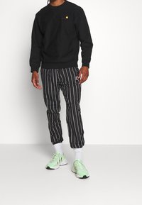 Musta pyöreäkauluksinen collegepaita, jossa on pieni logo, yhdistetty mustavalkoisiin raidallisiin housuihin. Päällä vaaleanvihreät urheilukengät ja valkoiset sukat.