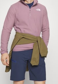 Fleece-pullover i lys lilla med halvlukket lynlås, logo på brystet. Olivengrøn jakke bundet om taljen, parret med marineblå shorts med lomme detalje.