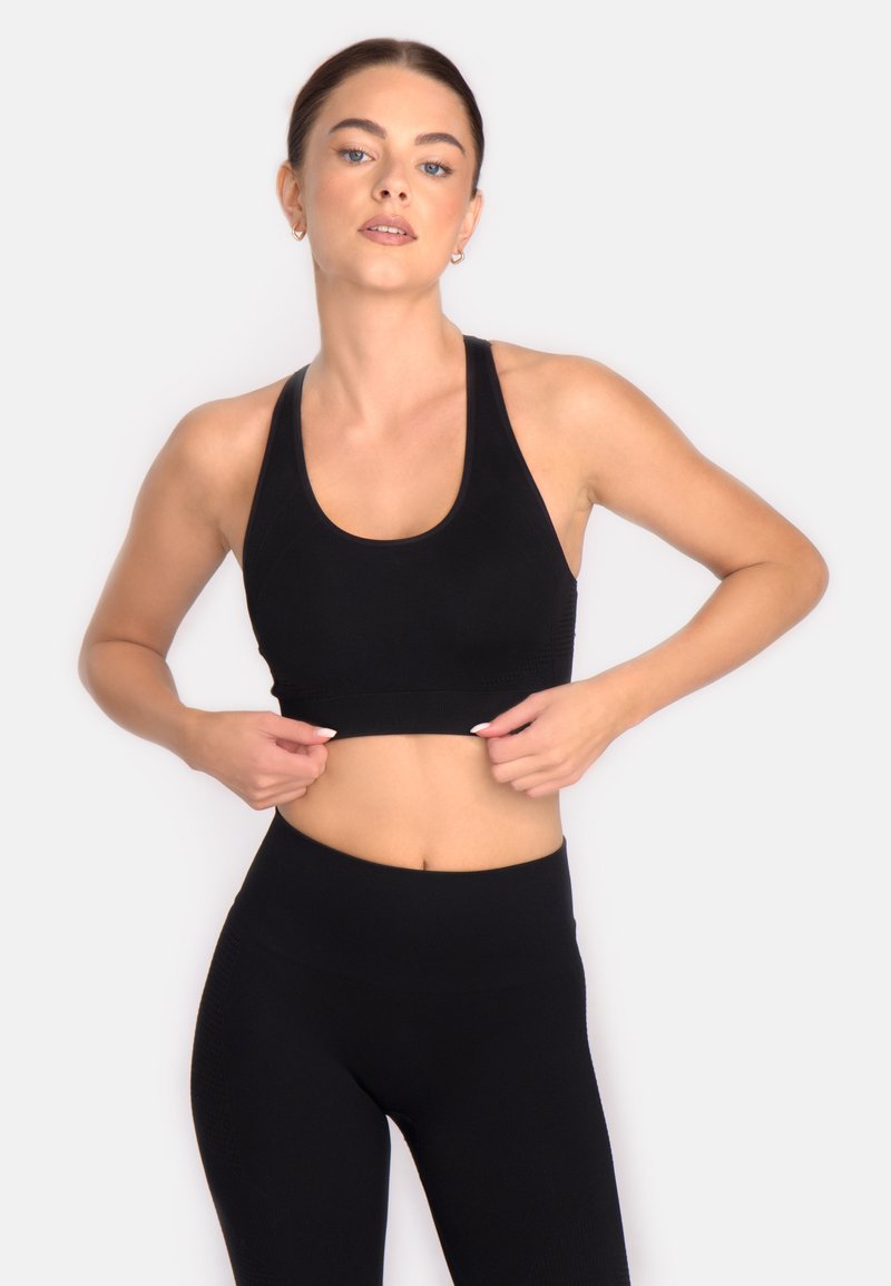 Sieviete, tērpusies melnā sporta krūšturī un legingos, pielāgo savu augšu pret neitrālu gaišu fona.