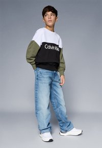 Sweatshirt em cores preto, branco e verde oliva com o logótipo "Calvin Klein", acompanhado de jeans baggy azuis claros e ténis brancos.