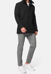 Black wool blend coat with button closure, high collar, and side pockets. Paired with gray slim-fit trousers and white sneakers.