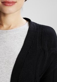 Cardigan nero lavorato a maglia con un motivo testurizzato, indossato sopra una maglietta grigia chiaro. Il cardigan presenta un ampio scollo a V e una chiusura aperta.