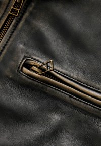 Zwart leer met een gestructureerde afwerking, voorzien van een bronzen ritssluiting en trekband, met gedetailleerde stiksels langs de randen.