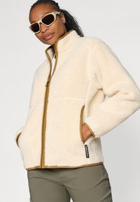 Crème-kleurige fleece jas met een hoge kraag, bruine ritssluiting en afwerking, en twee zijzakken. Gladde textuur met een zachte, warme uitstraling.
