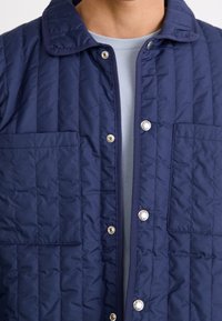Mörkblå quiltad jacka med knäppning framtill, två bröstfickor och krage. Slät textur med synlig sömnadsdesign och metallaccent.