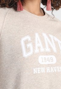 Sudadera de color beige claro con texto en blanco destacado "GANT 1949 NEW HAVEN." Tela suave con cuello redondo y acentos de borlas visibles.