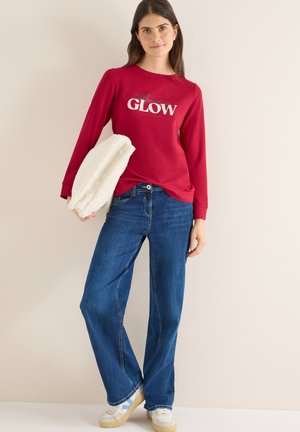 Czerwona koszula z długim rękawem z napisem "GLOW", zestawiona z niebieskimi spodniami typu wide-leg i białymi sneakersami. Modelka trzyma puszysty biały dodatek.