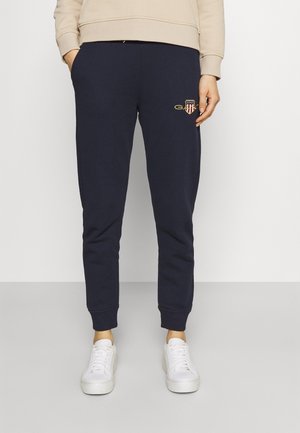 Personne portant un jogging bleu marine avec un petit emblème brodé sur la cuisse, un sweat-shirt beige et des baskets blanches, debout sur une surface grise.