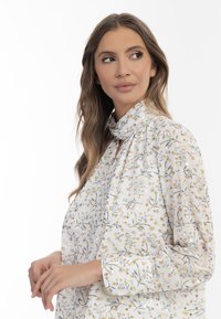 Langærmet bluse i lys creme med delikat blomsterdesign. Har en høj hals med sløjfe og en blød, flydende tekstur. Diskrete guld accenter.