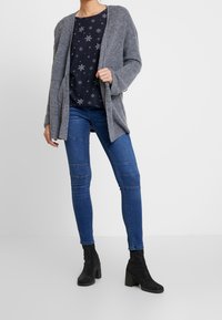 Cardigan gris en tricot porté sur un haut bleu marine orné de motifs flocons de neige blancs, associé à un jean bleu avec des accents cousus et des bottines noires à talons.