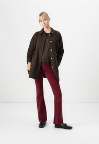 Donna in piedi con un cappotto buttonato marrone scuro, un maglione a treccia, pantaloni svasati bordeaux e mocassini neri, su uno sfondo bianco semplice.