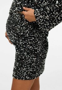 Zwangere persoon in een zwarte paillettenjurk, houdt de jurk met beide handen vast bij de ronde buik, zijaanzicht.