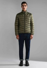Giacca a vento color verde oliva con colletto alto, caratterizzata da sezioni trapuntate orizzontali, chiusura con zip e tasche laterali; abbinata a jeans scuri.