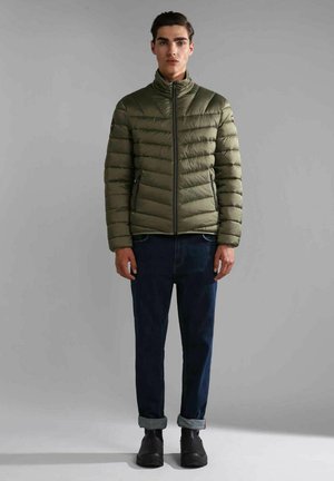 Giacca a vento color verde oliva con colletto alto, caratterizzata da sezioni trapuntate orizzontali, chiusura con zip e tasche laterali; abbinata a jeans scuri.