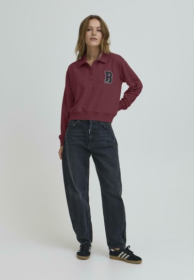 Bordó pulóver gallérral és hímzett 'B' betűvel a mellkasán, laza fazonú fekete farmernadrággal és fekete sportcipővel párosítva.