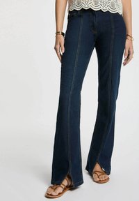 Jeans a zampa d'elefante blu scuro con fessure laterali, caratterizzati da cuciture a contrasto e una texture denim liscia, abbinati a sandali marroni con cinturini.