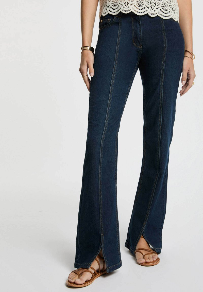 Jeans a zampa d'elefante blu scuro con fessure laterali, caratterizzati da cuciture a contrasto e una texture denim liscia, abbinati a sandali marroni con cinturini.