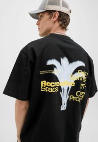 Zwart katoenen T-shirt met een grote witte palmboomafbeelding en gele tekst op de achterkant, korte mouwen en een ronde hals.