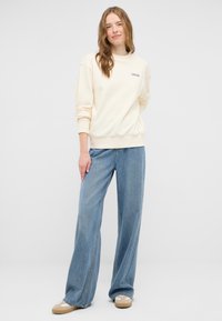 Felpa beige chiaro con polsini e orlo a coste, abbinata a jeans in denim blu chiaro a gamba larga. Calzature casual completano l'outfit.
