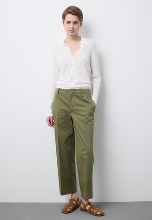 Jonge persoon met kort haar staat tegen een witte muur, draagt een wit shirt met lange mouwen, olijfgroene broek en bruine sandalen.