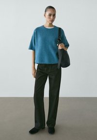 Vrouw draagt een kort mouwen trui in teal, zwarte brede broekspijpen, zwarte schoenen, en houdt een zwarte schoudertas vast tegen een effen achtergrond.