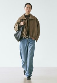 Frau, die eine braune Jacke über einem braunen Oberteil trägt, hellblaue Schlagjeans, schwarze Schuhe und eine große schwarze Umhängetasche bei sich hat.