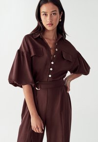 Camisa marrón de gran tamaño, de botones y con mangas abullonadas, con dos bolsillos en el pecho y cintura ceñida, con botones prominentes y una textura suave.