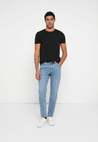 Camiseta negra de cuello redondo, vaqueros rectos de color azul claro, zapatillas blancas, modelo de pie con las manos en los bolsillos sobre un fondo liso.