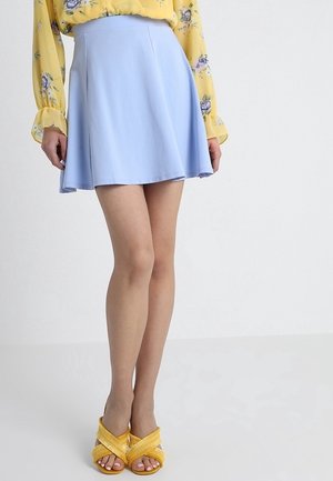 Femme portant une jupe évasée bleu clair, un chemisier jaune fleuri à manches longues et des sandales jaunes ouvertes à l’avant avec des lanières croisées.
