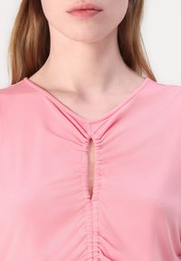 Rosa blus med rund halsringning, som har en nyckelhålöppning och ertad tygdetalj runt halsringningen. Slät, mjuk textur.