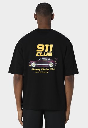 Homme portant un t-shirt noir avec le texte "911 Club" et un motif de voiture de course violette dans le dos sur un fond uni.