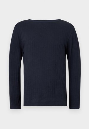 Rövid szőke hajú férfi, tengerészkék hosszú ujjú ingben, sötét farmerban és barna elegáns cipőben, egy kézzel a zsebében áll.