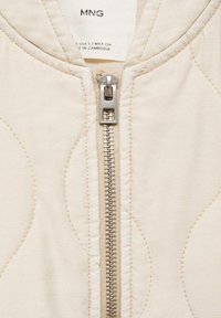 Beige quilted jacket with a metallic zipper and cream accents. Features smooth texture and circular stitching patterns. Label attached.