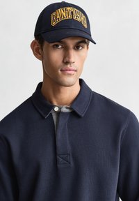 Marineblaue Mütze mit gelbem "GANT 1949" Schriftzug, getragen zu einem marineblauen Poloshirt mit grauem Kragen und einem dreieckigen Steppdetail an der Knopfleiste.