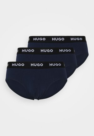 Trois paires de sous-vêtements de style slip bleu marine avec des ceintures élastiques noires présentant le logo "HUGO" en blanc. Texture lisse et design ajusté.