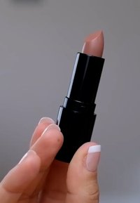 Pomadka w czarnej tubce, z zaokrąglonym, cielistym różowym wkładem. Ręka trzymająca ją ma neutralny wzór paznokci z białymi końcówkami.