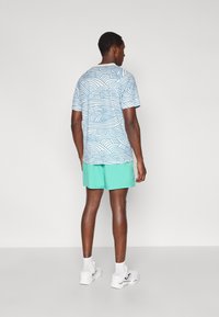 Μπλε και λευκό t-shirt με σχέδιο κυμάτων, κοντό πράσινο σορτς, λευκά αθλητικά παπούτσια, στέκεται από πίσω με κοντά μανίκια και στρογγυλή λαιμόκοψη.