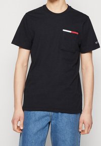 T-shirt en coton noir à manches courtes, col rond, et une poche plate avec des rayures horizontales rouges et blanches sur la ligne de poche.