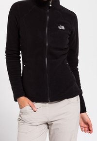 Musta fleece-takki, jossa on korkea kaulus, vetoketjukiinnitys ja sivutaskut. Vasemmassa rinnassa on logo. Pehmeä rakenne, istuva malli.