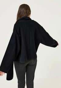 Giacca nera oversize con maniche larghe, morbida al tatto e colletto alto. I dettagli delle cuciture lungo la schiena aggiungono struttura. Indossata con jeans scuri.