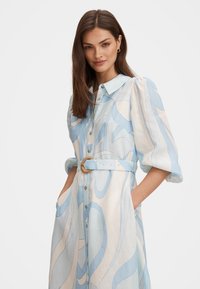 Femeie purtând o rochie cu mâneci lungi, cu centură, cu modele turbinante albastre și bej, stând cu mâinile în buzunare, pe un fundal simplu.