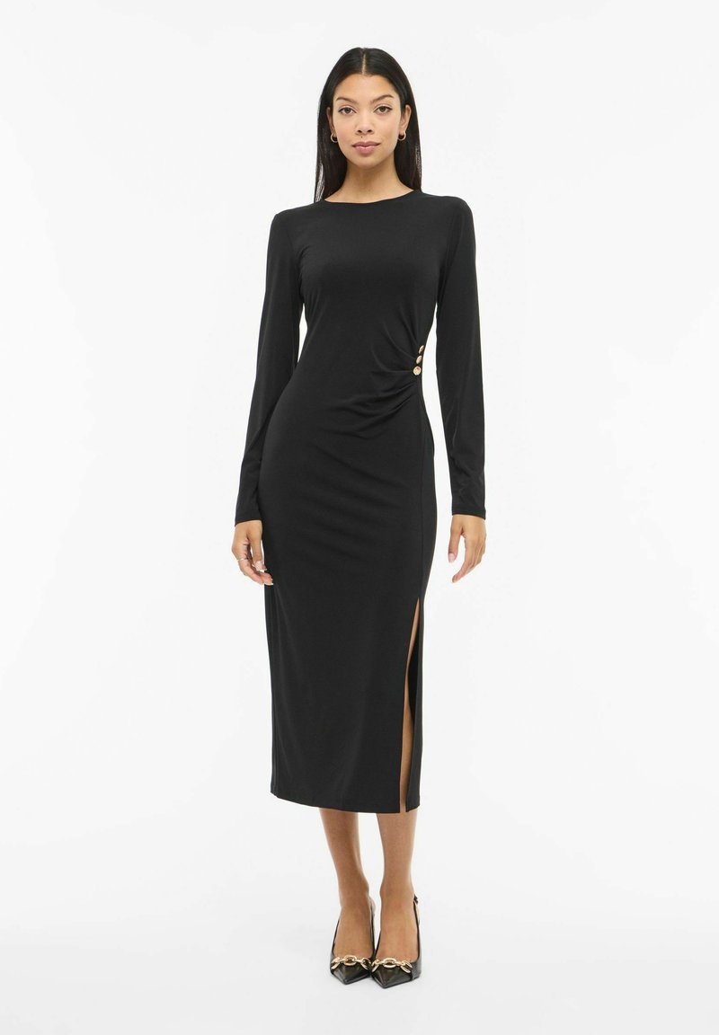 Juoda ilgomis rankovėmis suknelė su šoniniu skeltuku, drapiruotu dizainu ir trimis auksinės spalvos mygtukais juosmens akcentui. Minkšta, lygi audinio tekstūra.