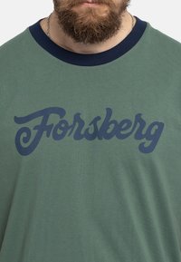 Camiseta verde de manga corta con un cuello acanalado en azul marino y un gran texto cursivo azul que dice "Forsberg" en el frente. Textura de tela suave.