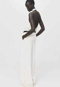 Mono blanco con cuello halter y espalda descubierta, piernas amplias y un cinturón negro en la cintura. Tejido suave con un ligero brillo.