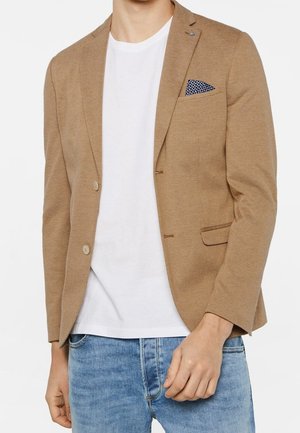 Man draagt een lichtbruine blazer met textuur, een marineblauw zakdoekje met patroon, een wit T-shirt met ronde hals en lichtblauwe spijkerbroek.